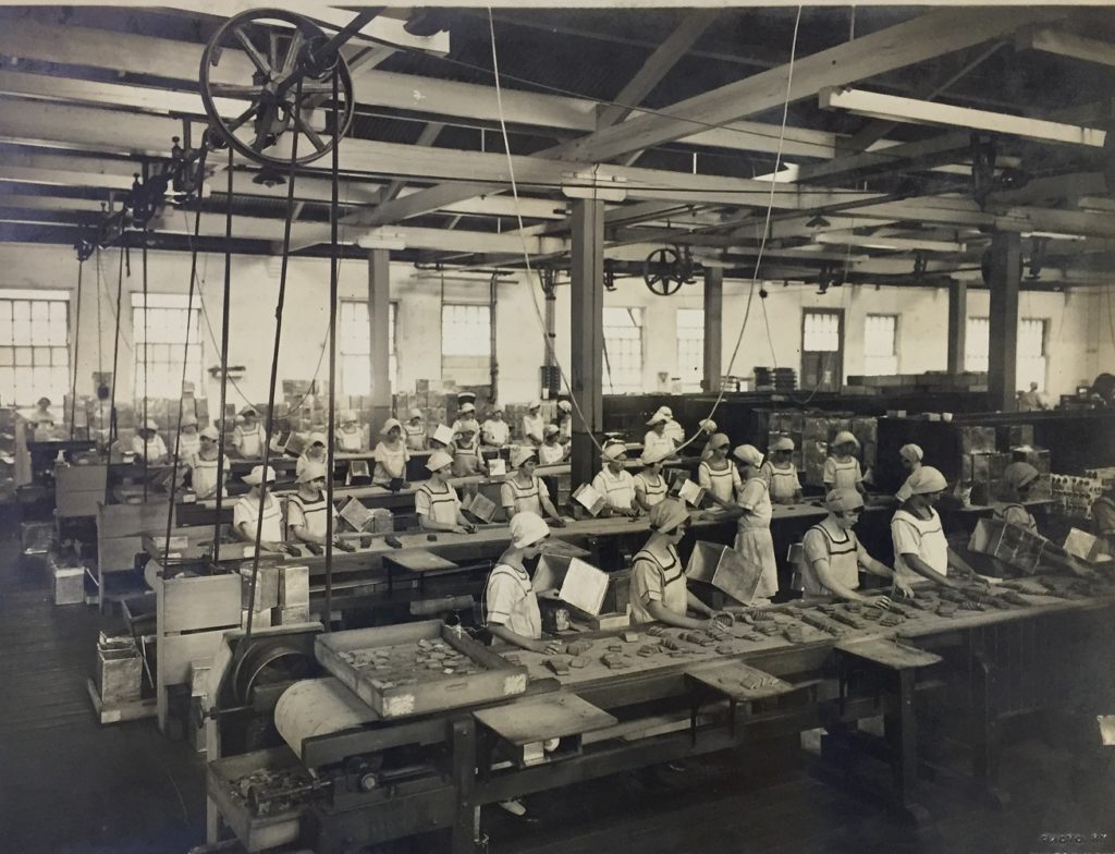 Black and white photograph of a group of female factory workers standing before four long conveyor belts packing biscuits into metal boxes. There are approximately ten women per conveyor belt. 